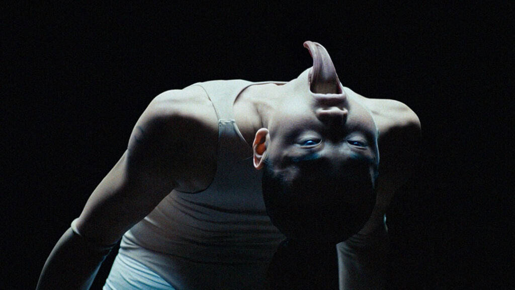 Contortionist dancer in Bonnie Banane’s ‘Hoes of Na’ bending backward in a dark, surreal setting with an elongated tongue effect.