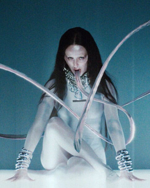 Bonnie Banane depicted with elongated, ribbon-like tongues in a futuristic blue setting from her video ‘Hoes of Na’.