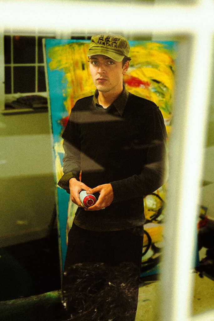 Studio portrait of artist Tomas Gittins holding a spray can in front of a large abstract painting
