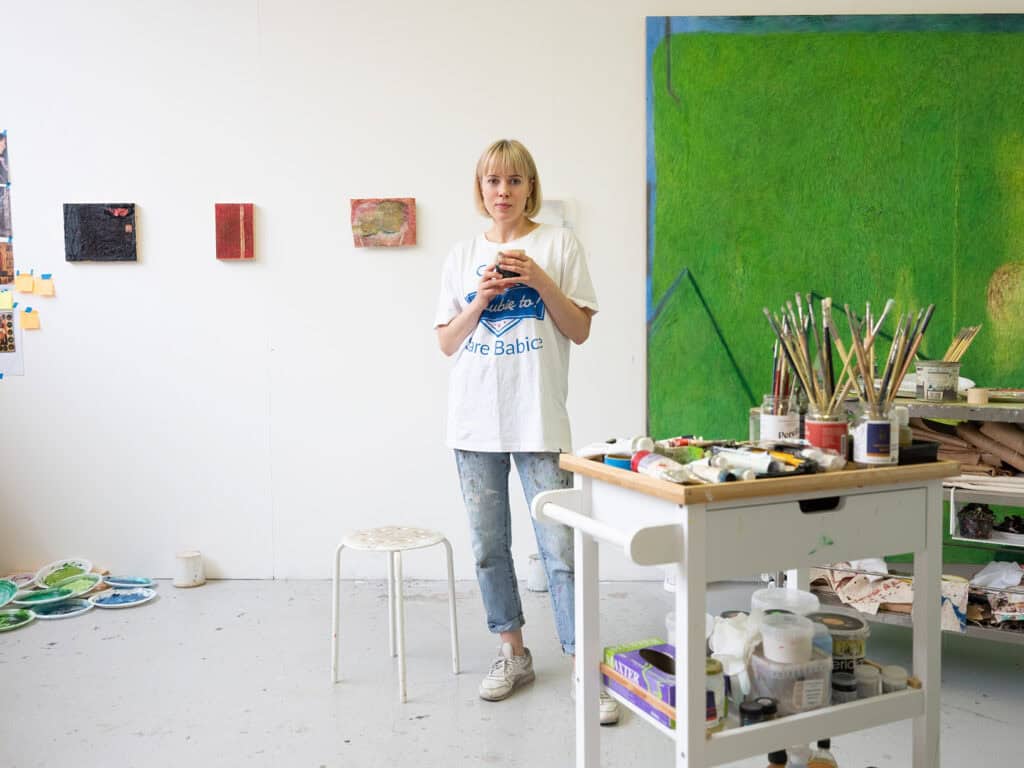 Photograph of artist Karolina Albricht in her studio, surrounded by paint tubes, brushes, and abstract canvases in progress.