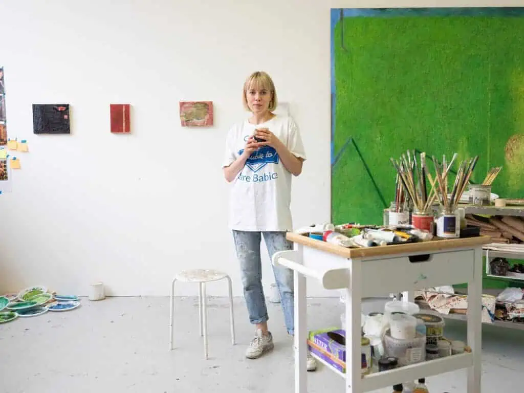 Photograph of artist Karolina Albricht in her studio, surrounded by paint tubes, brushes, and abstract canvases in progress.