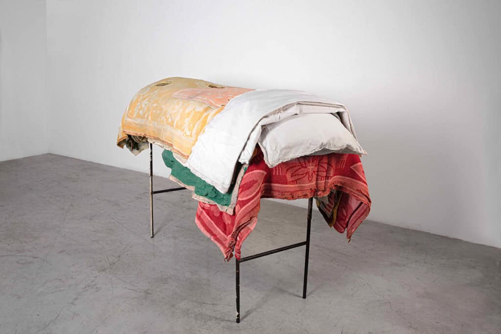 Berlinde De Buyckere’s installation at Galleria Continua during Frieze Masters 2025, featuring a cot-like structure draped in layered blankets exploring themes of warmth, fragility, and human vulnerability.