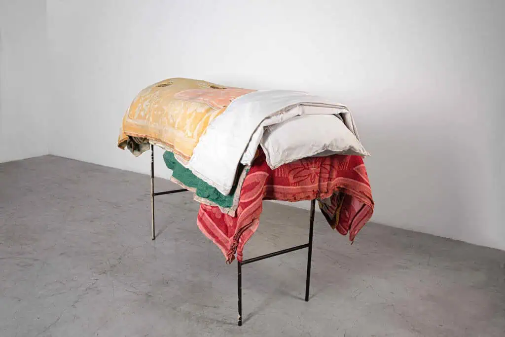Berlinde De Buyckere’s installation at Galleria Continua during Frieze Masters 2025, featuring a cot-like structure draped in layered blankets exploring themes of warmth, fragility, and human vulnerability.