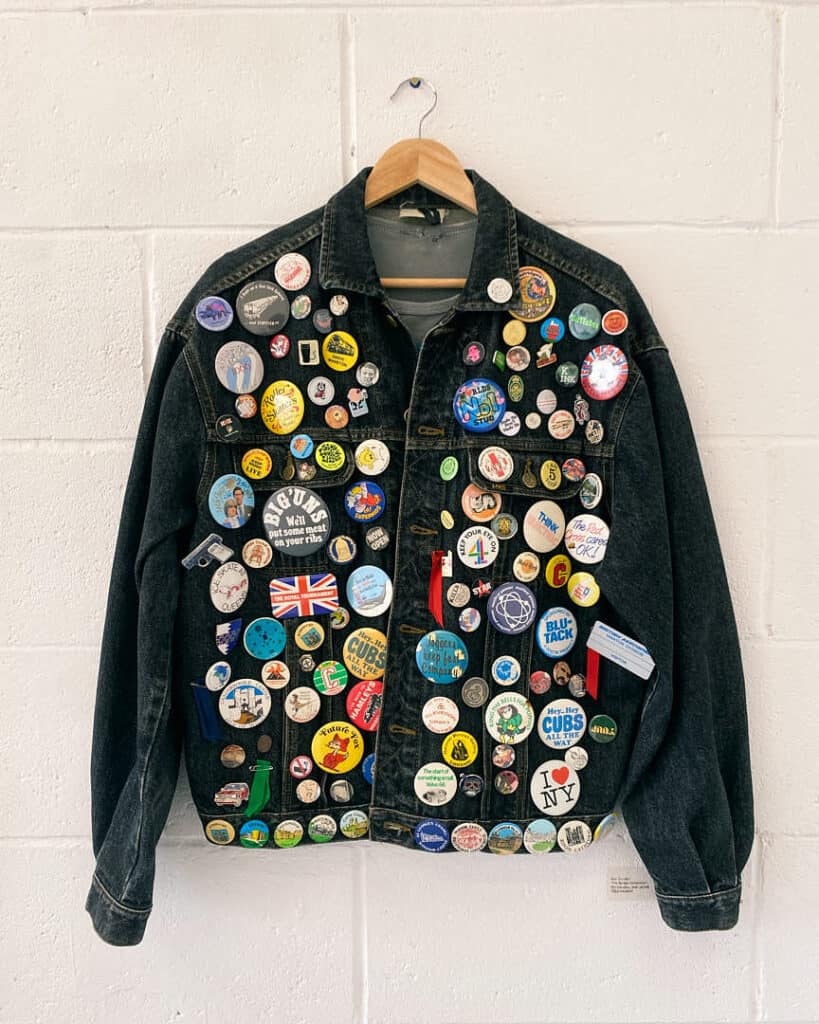 Denim jacket covered in colorful collectible badges displayed on a white wall.