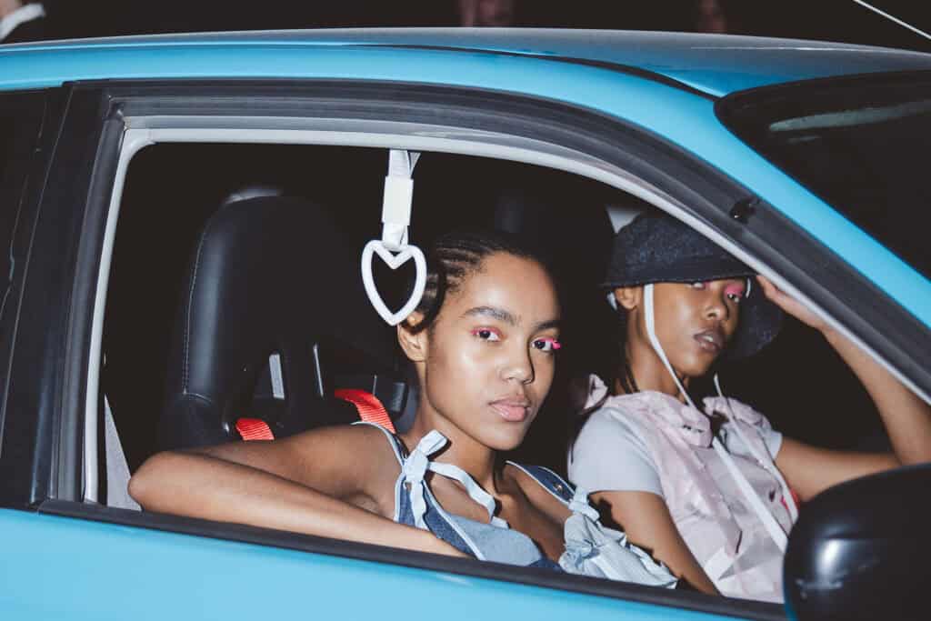 Two models sitting inside a blue car backstage at Florentina Leitner SS26 show, featuring playful pink eye makeup and early-2000s inspired outfits.