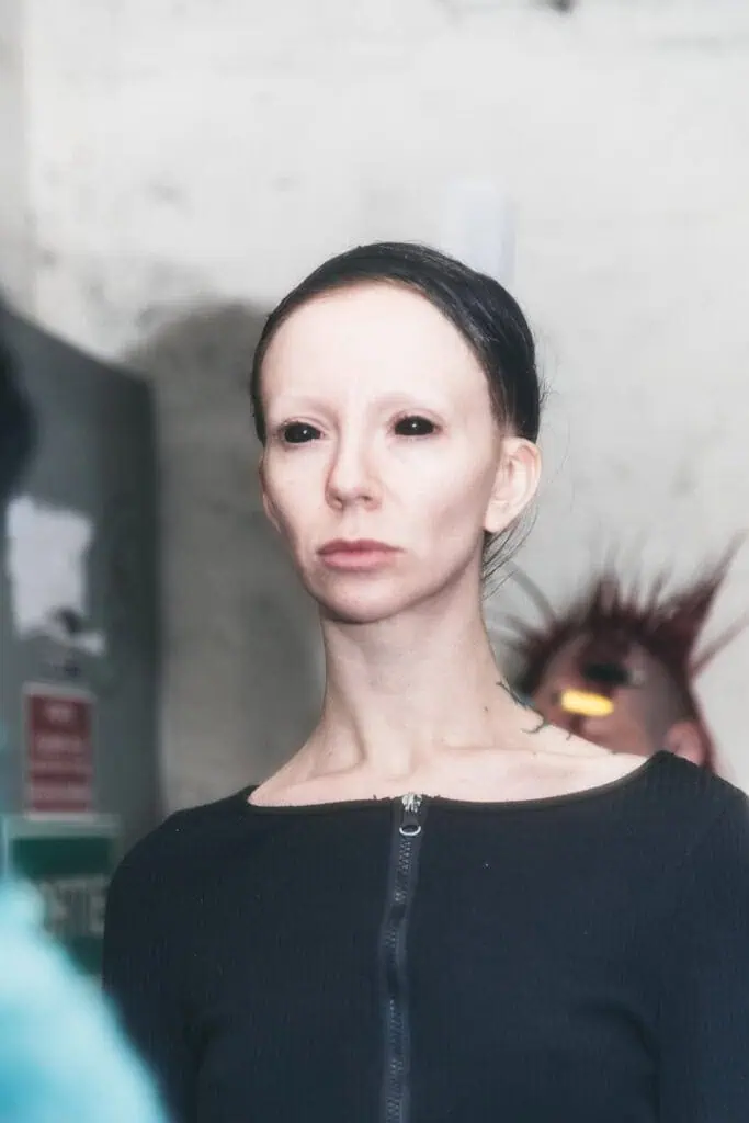 Close-up of model with black-out lenses and bleached brows in industrial fashion setting.