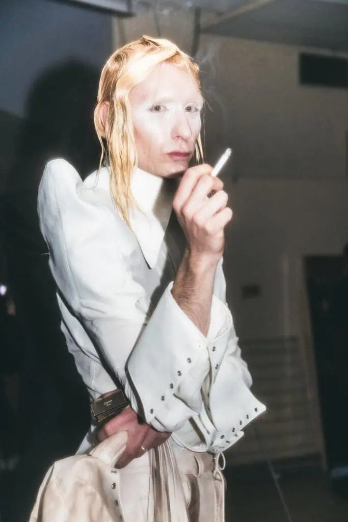 Model in structured all-white look with ethereal makeup smoking backstage.