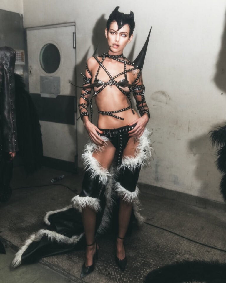 Model in dark couture look with leather, spikes, and faux fur in backstage setting.
