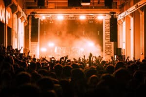 A large crowd in front of the Linecheck stage, illuminated by warm orange lights, with people raising their hands during a live concert.