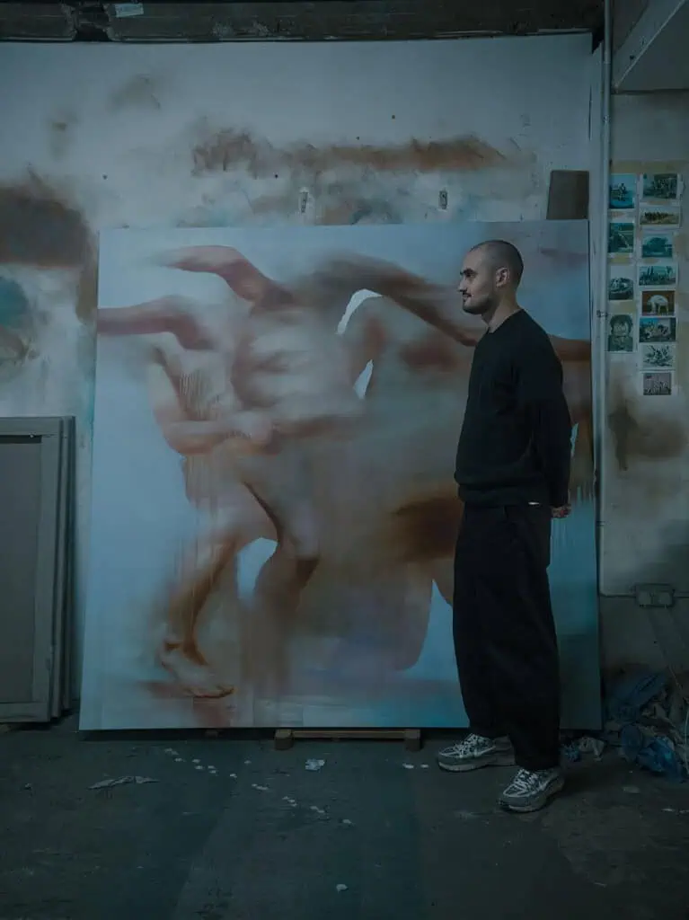 Photograph of artist Preslav Kostov standing beside his large-scale painting in a dimly lit studio, surrounded by sketches and materials.