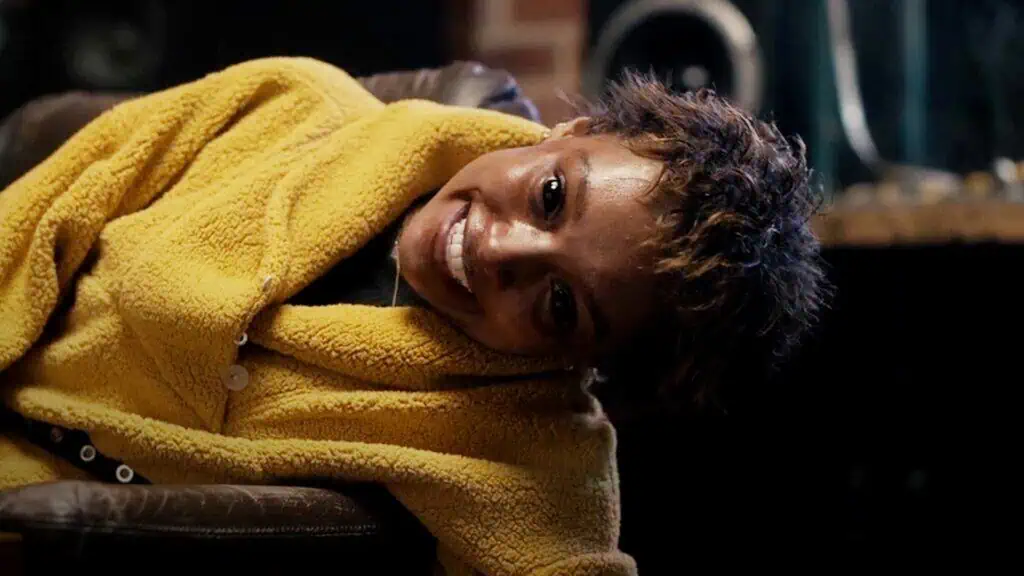 Scene from The Together (2025) showing character in yellow coat smiling on chair, part of Coralie Fargeat’s body horror film.