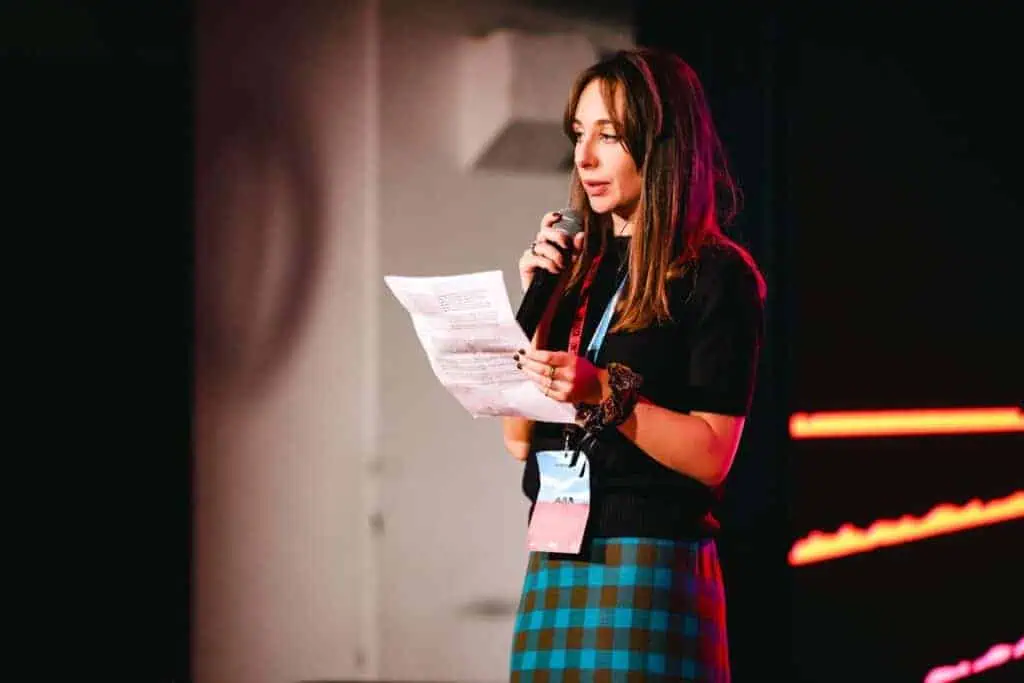 Anna Zò speaking on stage at Linecheck, holding a microphone and a printed sheet, with red stage lights behind her.