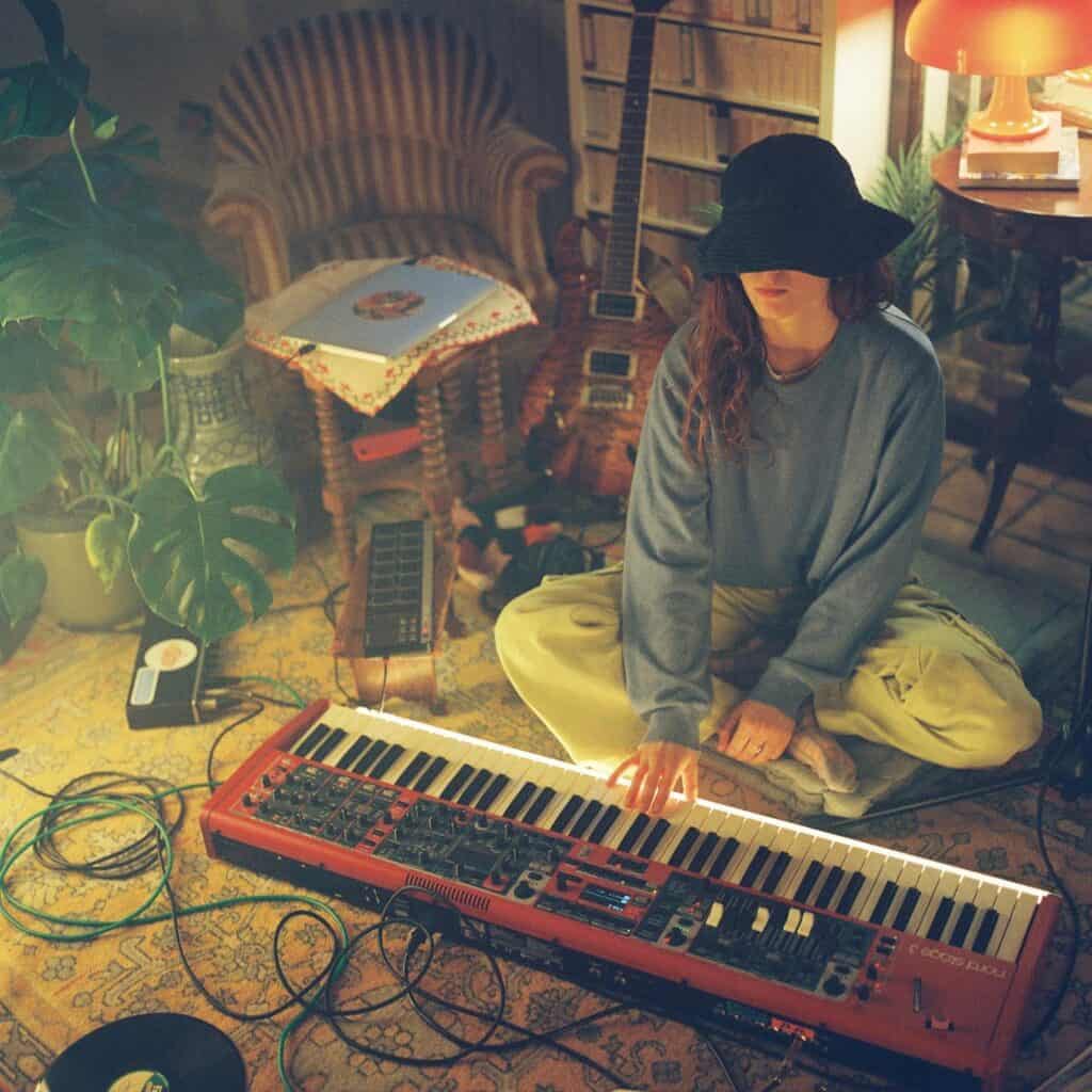Coca Puma sits cross-legged on the floor of a cozy, dimly lit room, playing a red keyboard surrounded by instruments, cables, vinyls, and warm ambient light.