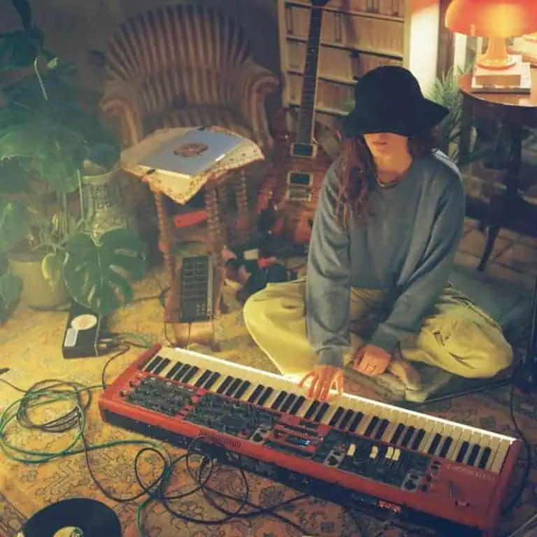 Coca Puma sits cross-legged on the floor of a cozy, dimly lit room, playing a red keyboard surrounded by instruments, cables, vinyls, and warm ambient light.