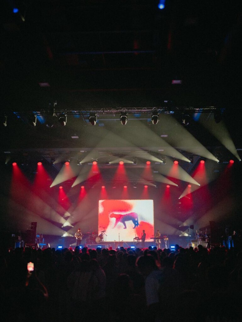 A live concert by Coca Puma, with the stage lit in red and yellow beams and a central screen showing an abstract moving figure as the audience watches.
