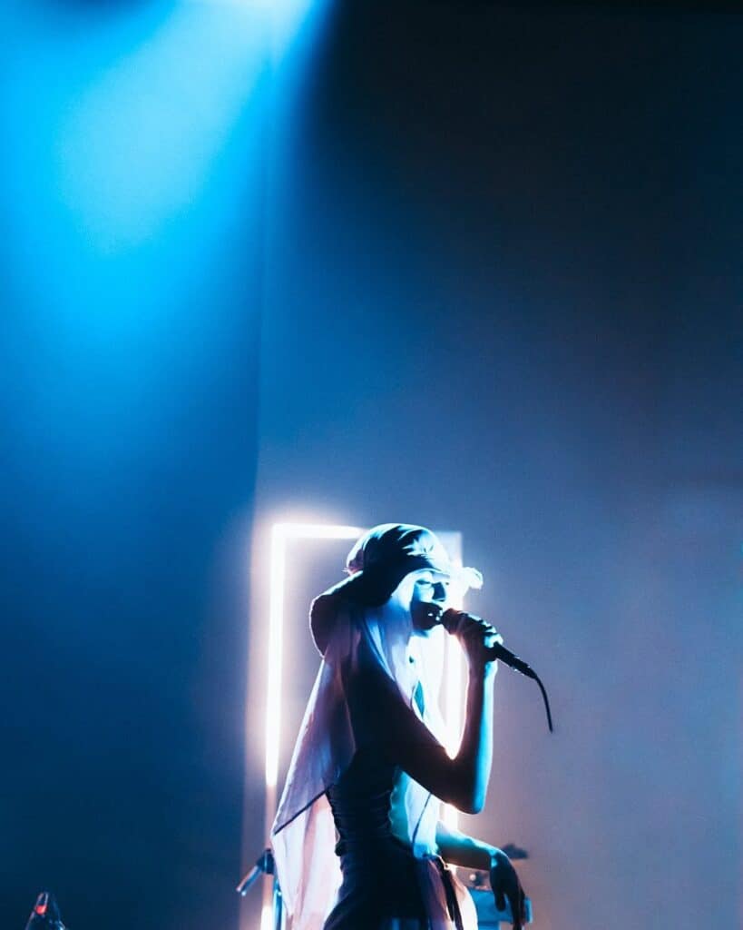 Coca Puma performs under soft blue stage lighting, singing into a microphone while wearing a draped headpiece in front of a glowing rectangular frame.