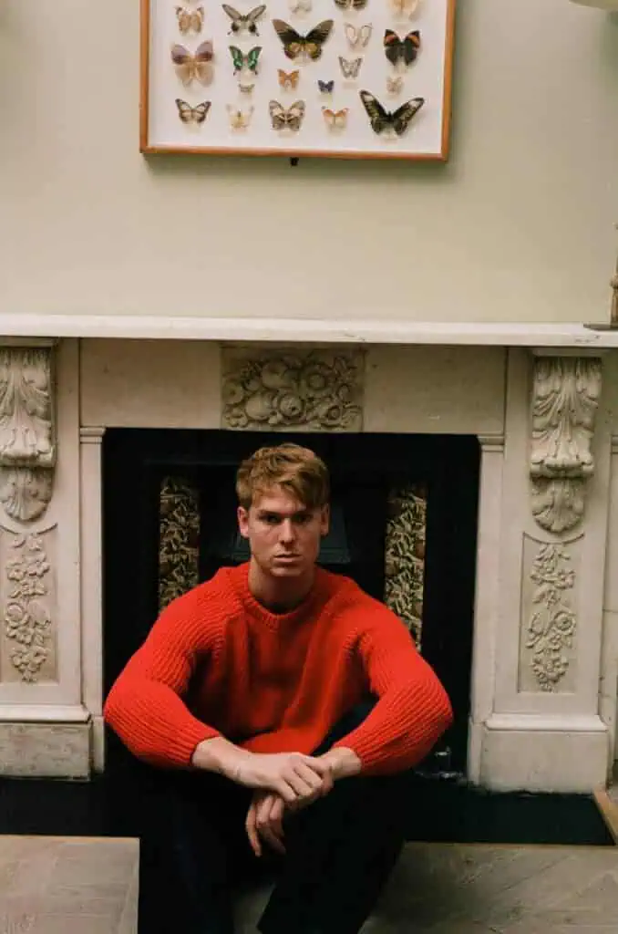Dan Whitlam, a man with short hair in a bright red sweater, sits on the floor in front of an ornate white fireplace, with a framed display of butterflies hanging above the mantel.