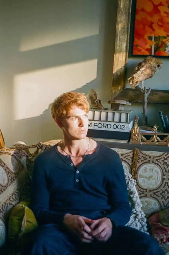 Dan Whitlam sits on a patterned sofa in a sunlit room, looking to the side—behind him are decorative objects, a framed painting, and a lightbox sign reading "JIM FORD 002," evoking the cool style of Dan Whitlam.
