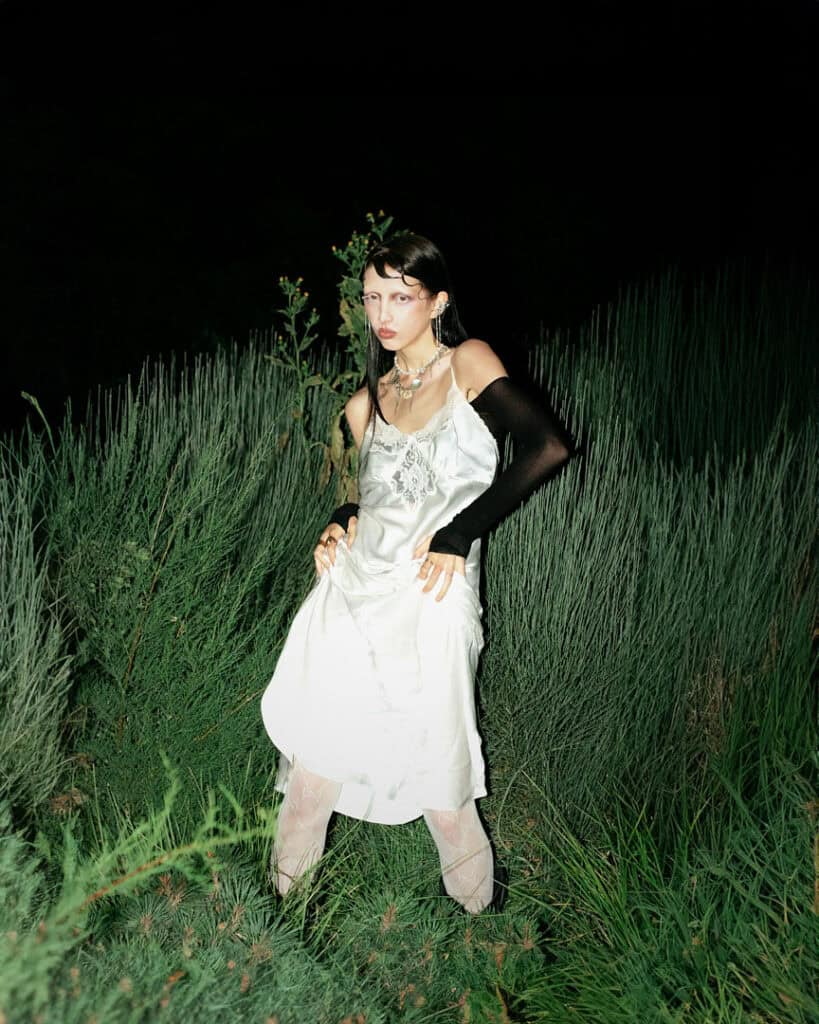 A woman in a white dress and long black glove poses amid tall green grass at night. Dramatic Veronika Butkevich photography highlights her headpiece, statement necklace, and sheer stockings against the dark background.
