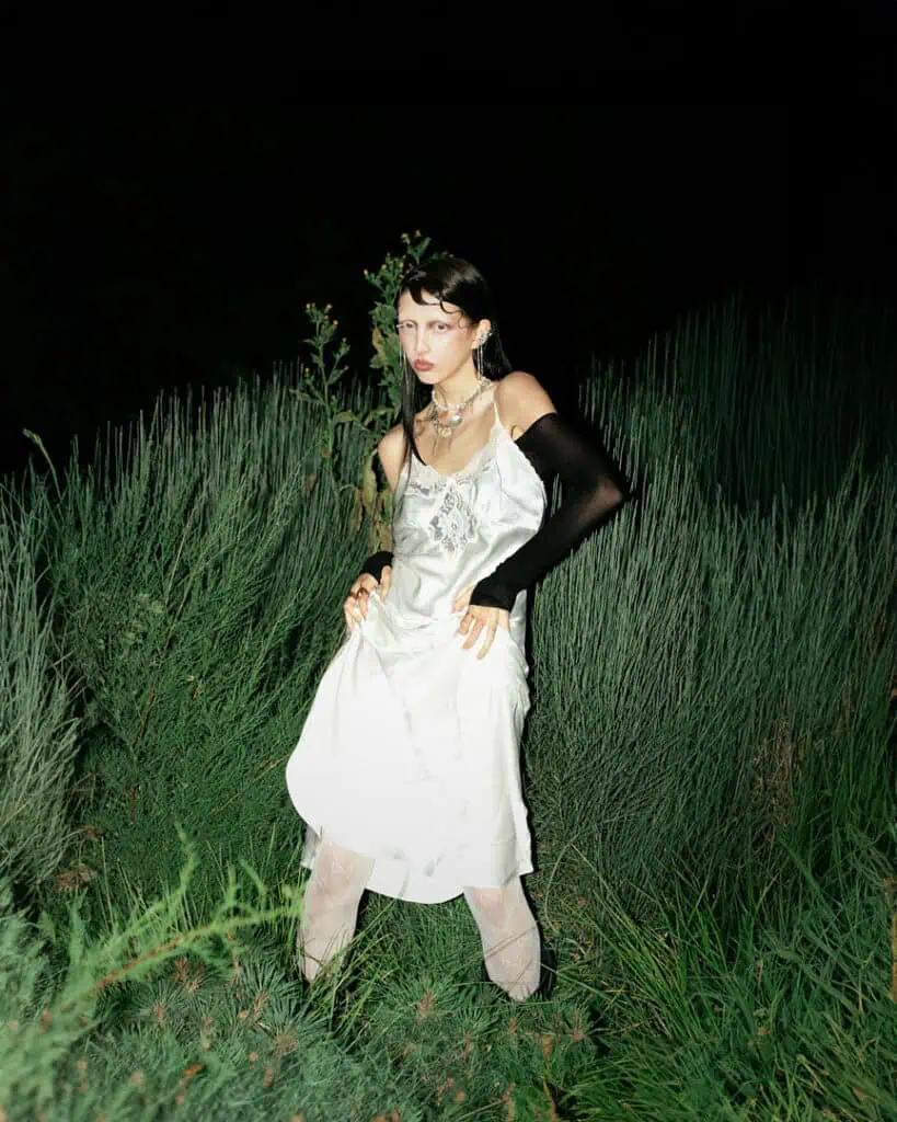 A woman in a white dress and long black glove poses amid tall green grass at night. Dramatic Veronika Butkevich photography highlights her headpiece, statement necklace, and sheer stockings against the dark background.