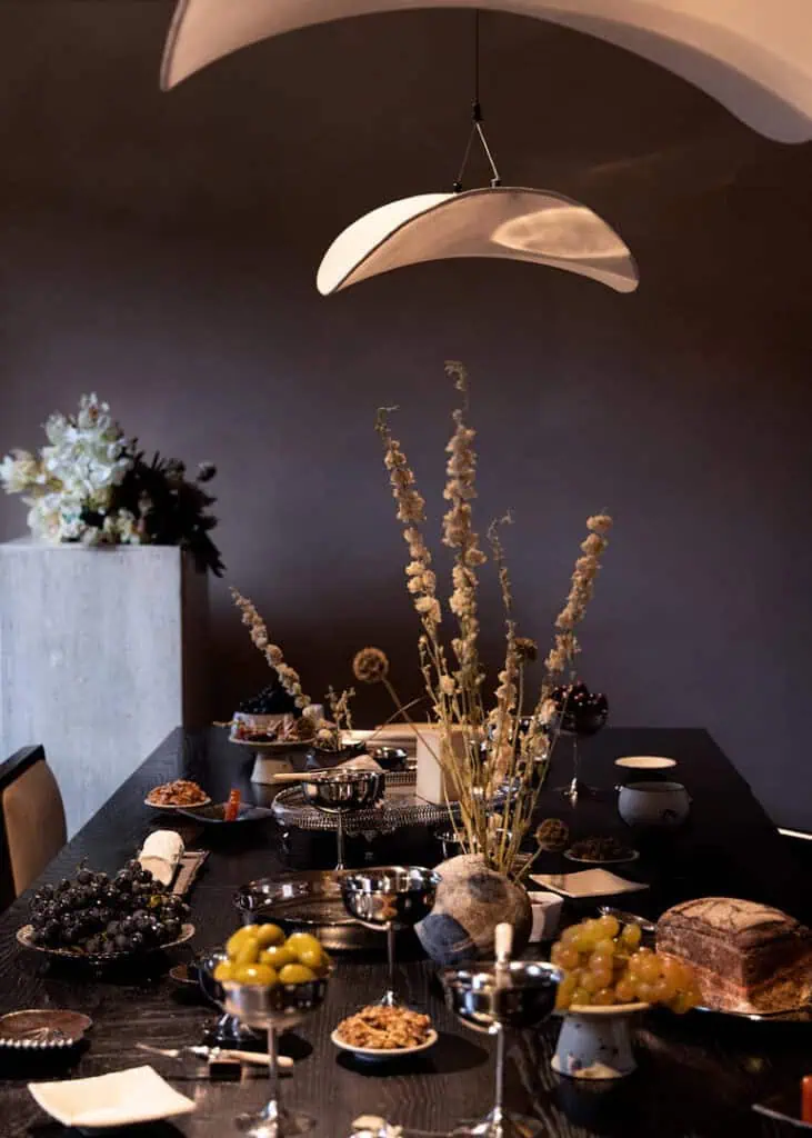A stylish dining table is set with fruit, bread, and elegant dishes under a modern white pendant light at the Mira Maktabi and Studio Deïmé event. Dried floral arrangements and soft lighting create a cozy, sophisticated atmosphere.
