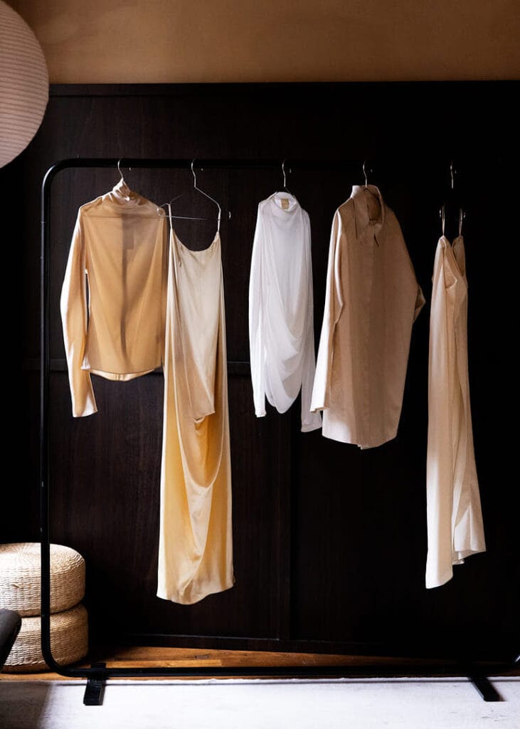 Five neutral-colored garments, including blouses and draped dresses, hang neatly on a black clothing rack in a dimly lit room with dark wood paneling and woven baskets on the floor at the Mira Maktabi and Studio Deïmé event.