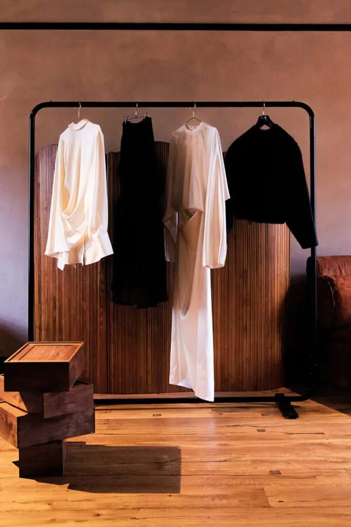 A clothing rack displays four garments—two white, one black, and one transparent black—hanging in front of a wooden room divider at Mira Maktabi and Studio Deïmé event. Wooden stacked boxes and part of a brown sofa rest on a wooden floor.