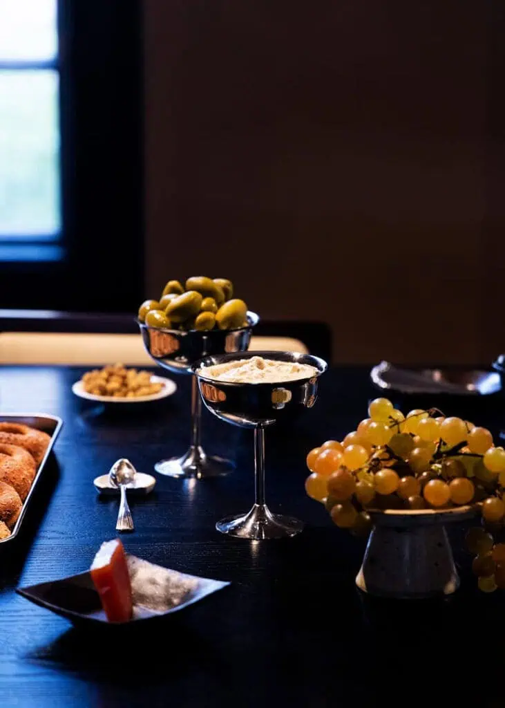 A dark table at Mira Maktabi and Studio Deïmé event is set with dishes of green olives, grapes, nuts, and dips in glass bowls, accompanied by pastries and a slice of grapefruit, all softly lit by natural light from a window.