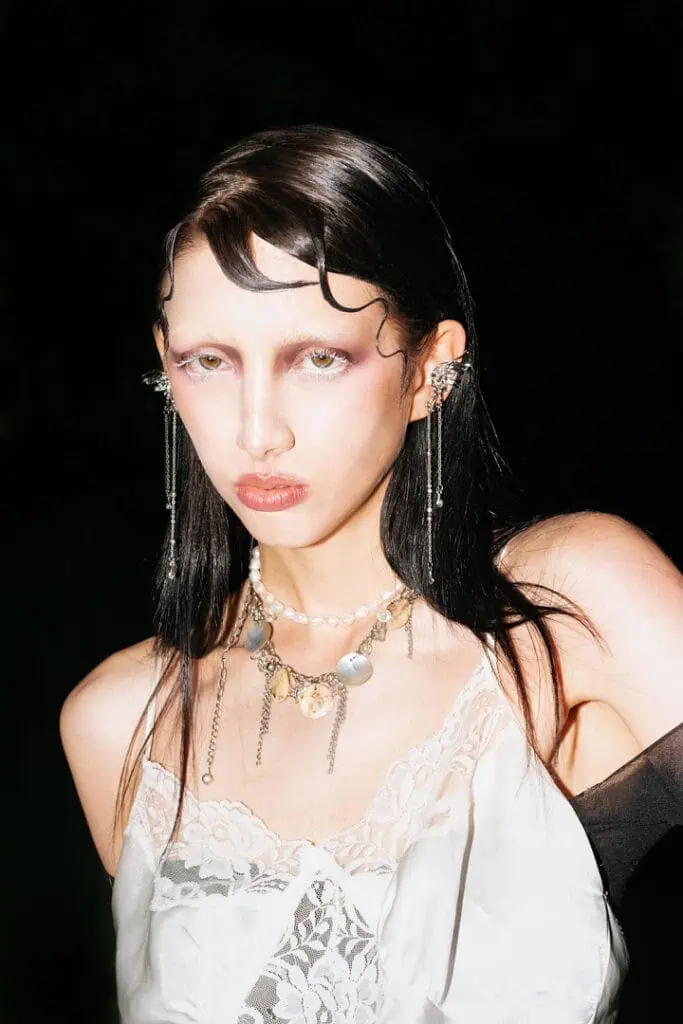 A person with slicked-back dark hair, bold makeup, and light eyebrows wears layered necklaces and dangling earrings with a white lace top, posing confidently against a dark background in the style of Veronika Butkevich photography.