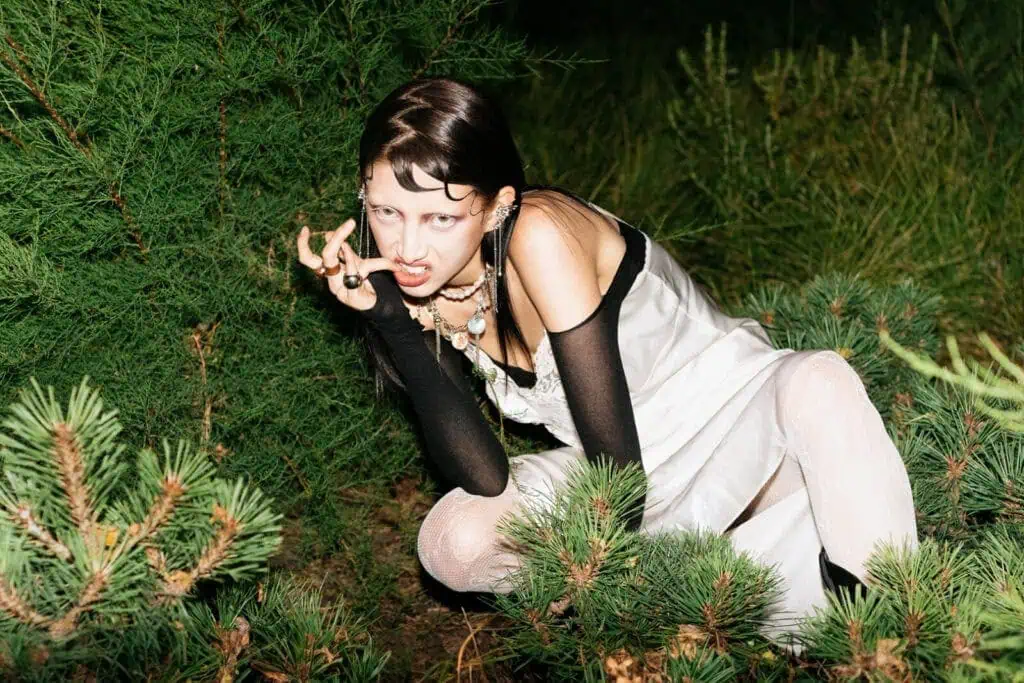 A person in a white dress with black sleeves crouches amid green bushes at night, baring their teeth and making a claw-like gesture with one hand—a striking, wild expression captured by Veronika Butkevich photography.