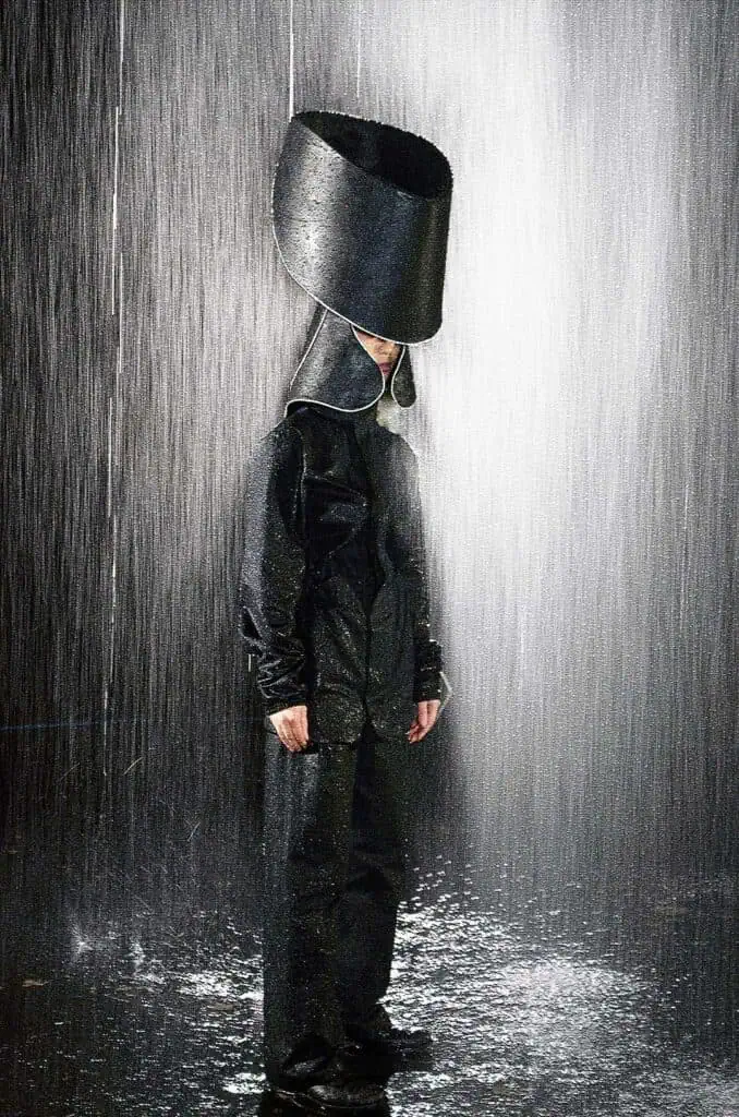 A person wearing a large, black, bucket-shaped hat designed by MIJODA DAJOMI and a shiny black outfit stands under heavy, pouring water—channeling bold style like the 5 brands you should know—with water cascading dramatically around them.