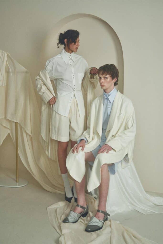 Two people in modern, minimalist cream and white outfits pose in a soft, neutral-toned room. Draped fabrics and curved shapes add sophistication to the serene setting. Everything designed by ZIMISM