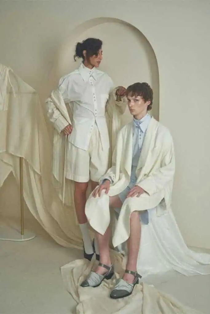Two people in modern, minimalist cream and white outfits pose in a soft, neutral-toned room. Draped fabrics and curved shapes add sophistication to the serene setting. Everything designed by ZIMISM
