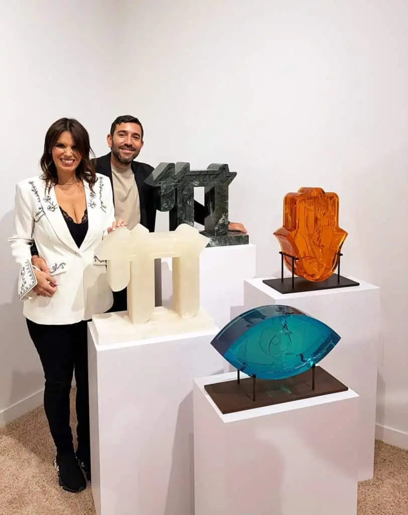 Two artists stand smiling beside four colorful sculptures displayed on white pedestals in an art gallery during Miami Art Week 2025. The sculptures include abstract shapes in white, blue, orange, and dark green.