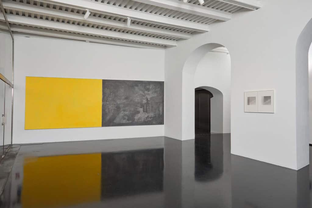 A modern art gallery with white walls and a dark shiny floor features works from the Atto 2/3: Col Tempo exhibition, including a large painting split between yellow and dark gray, two smaller framed pieces, high ceilings, and arched doorways.
