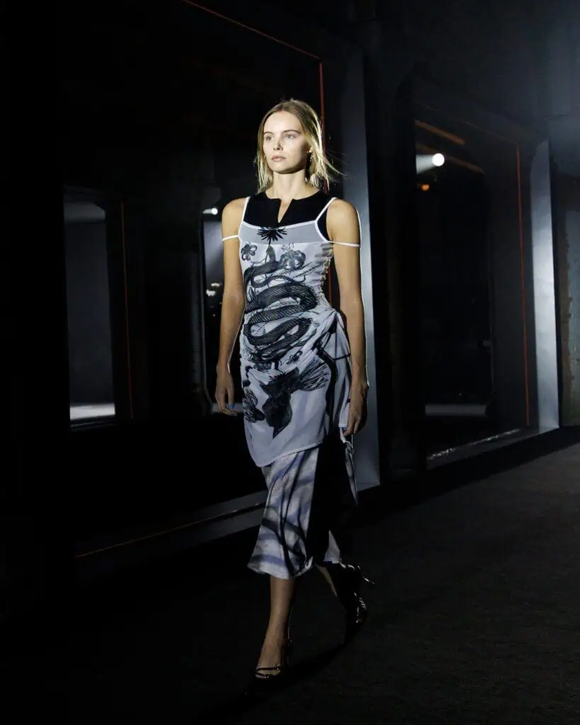 A model walks down a dimly lit runway wearing a sleeveless, black and white dress designed by ACCEPTANCE LETTER designer, with abstract patterns—a look inspired by the artistry of the 5 brands you should know. The background features dark walls and dramatic lighting.