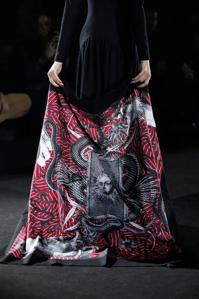 A person in a black dress designed by ACCEPTACE LETTER holds out the hem to showcase striking red, black, and white patterns featuring snakes and a detailed feline face at the center.