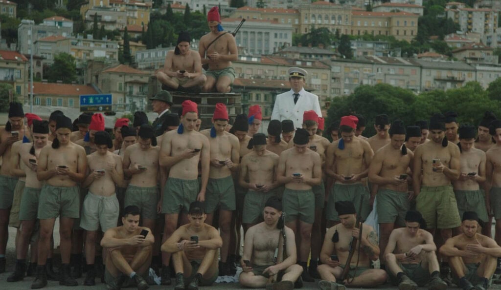 A large group of young men in shorts, many shirtless and wearing red or black caps, stand closely together outdoors, most looking down at their smartphones. A man in a white naval uniform stands among them. This is a still from ‘Fiume o Morte!’ by Director Igor Bezinović.