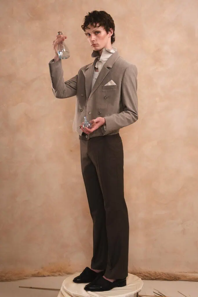A person in a gray suit stands on a round pedestal, holding a clear glass perfume bottle aloft in one hand and another bottle in the other, posing against a soft beige background. Everything designed by ZIMISM.
