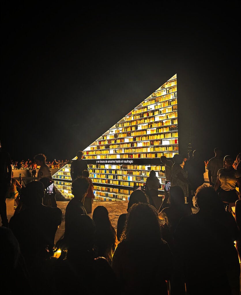 A crowd gathers at night around a large, triangular illuminated bookshelf filled with books, glowing brightly against the dark sky during Miami Art Week 2025.