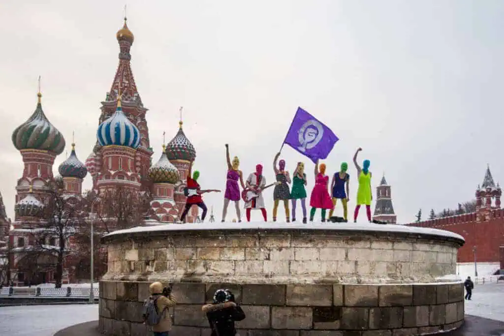 Maria Alyokhina of Pussy Riot and others, dressed in colorful outfits and masks, stand on a stone platform in Red Square, Moscow, holding a purple flag. St. Basil's Cathedral and the Kremlin loom behind them in the snow as two photographers capture the scene below.