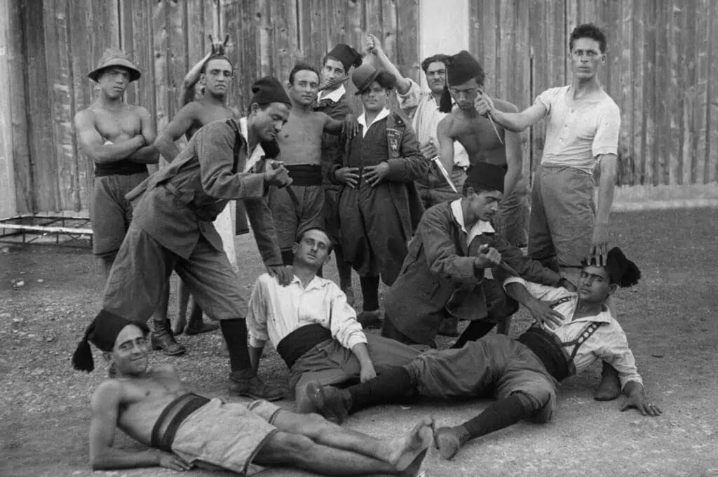A group of men in traditional clothing and fezzes pose playfully outdoors in front of a wooden fence, some standing, others sitting or lying on the ground—a relaxed, joyful still from ‘Fiume o Morte!’ by Director Igor Bezinović.