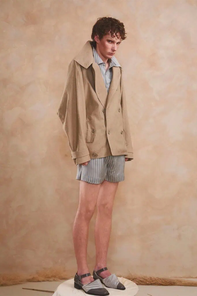 A young man stands on a small pedestal wearing an oversized beige blazer over blue and white striped shorts and shirt, styled with black-and-grey shoes, pink socks. Everything designed by ZIMISM.