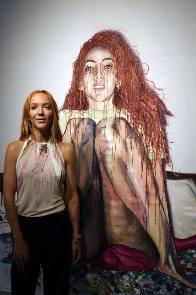 A woman with long red hair stands smiling in front of a large, detailed painting of herself at Miami Art Week 2025, both sporting similar red hair. The background is mostly white with colorful accents near the bottom.