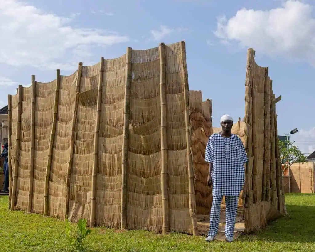 A person in patterned clothing stands before a structure of wooden poles and woven thatch at The Black Muse Art Festival 2025, set on a grassy area beneath a partly cloudy sky.
