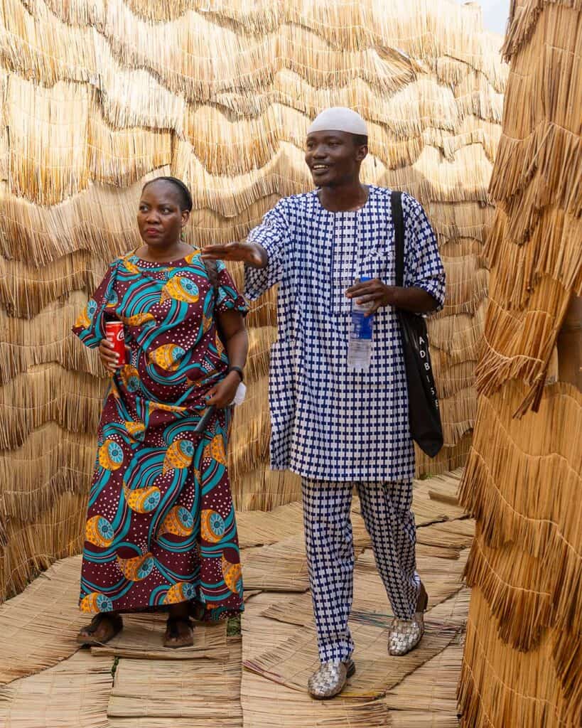 A woman and a man walk together on a woven mat path between tall straw walls at The Black Muse Art Festival 2025. She wears a colorful patterned dress and holds a drink; he wears patterns too, carries a bag, and points ahead while smiling.