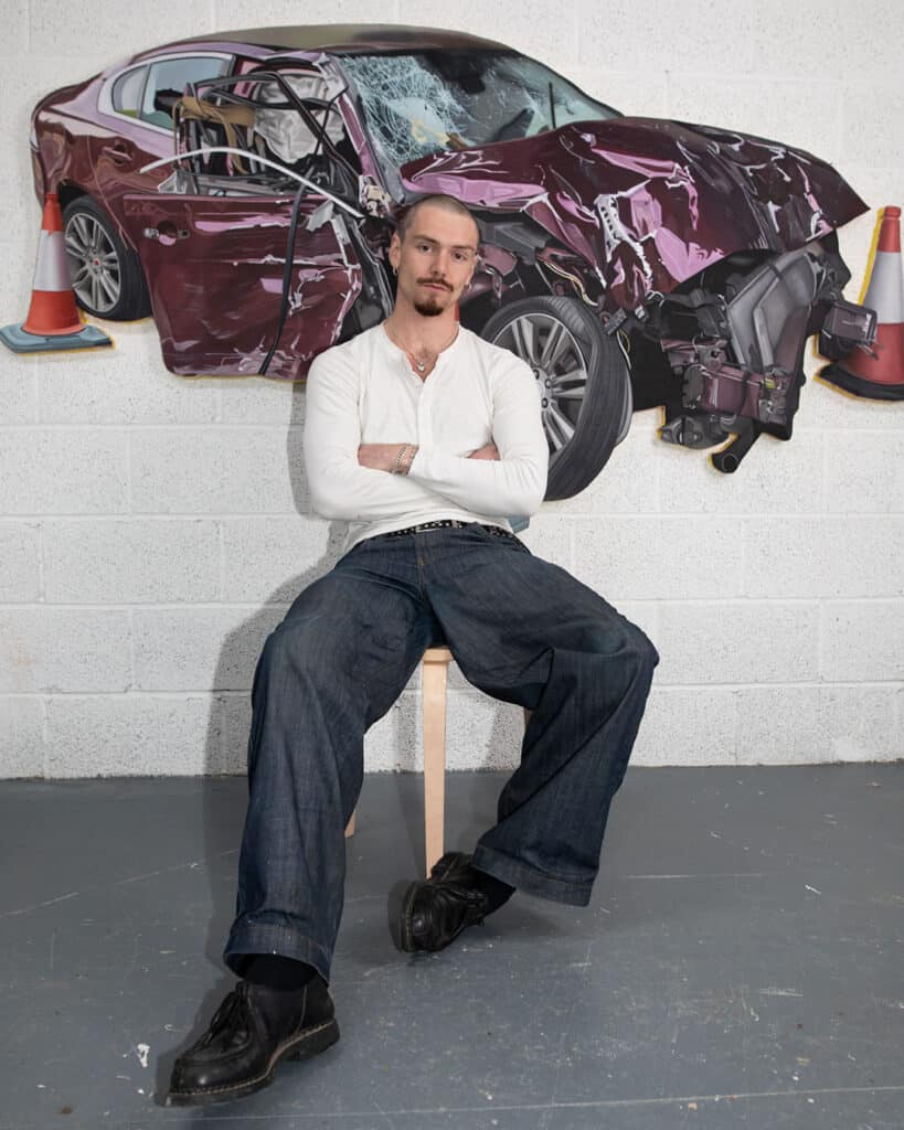 Callum Eaton, a man with a goatee wearing a white long-sleeve shirt and baggy jeans, sits on a stool with arms crossed in front of wall art depicting a severely crashed car.