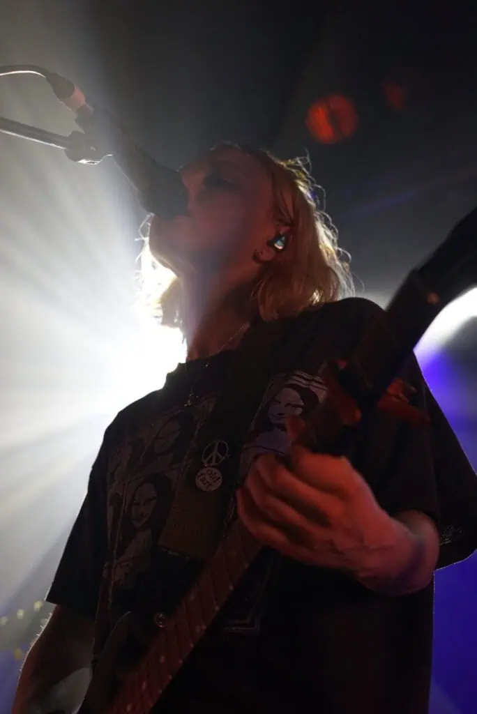 A musician with shoulder-length blonde hair from the Momma band sings into a microphone and plays electric guitar on stage, illuminated by dramatic backlighting that creates a striking silhouette effect.