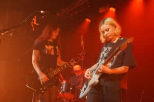 Two musicians from the Momma band play electric guitars on stage under orange lights, with a drummer in the background. Both guitarists wear black T-shirts and display their tattoos, creating an energetic and focused atmosphere.