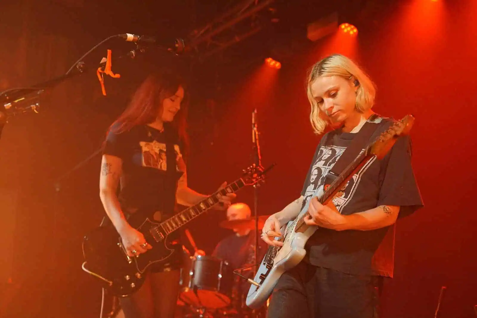 Two musicians from the Momma band play electric guitars on stage under orange lights, with a drummer in the background. Both guitarists wear black T-shirts and display their tattoos, creating an energetic and focused atmosphere.